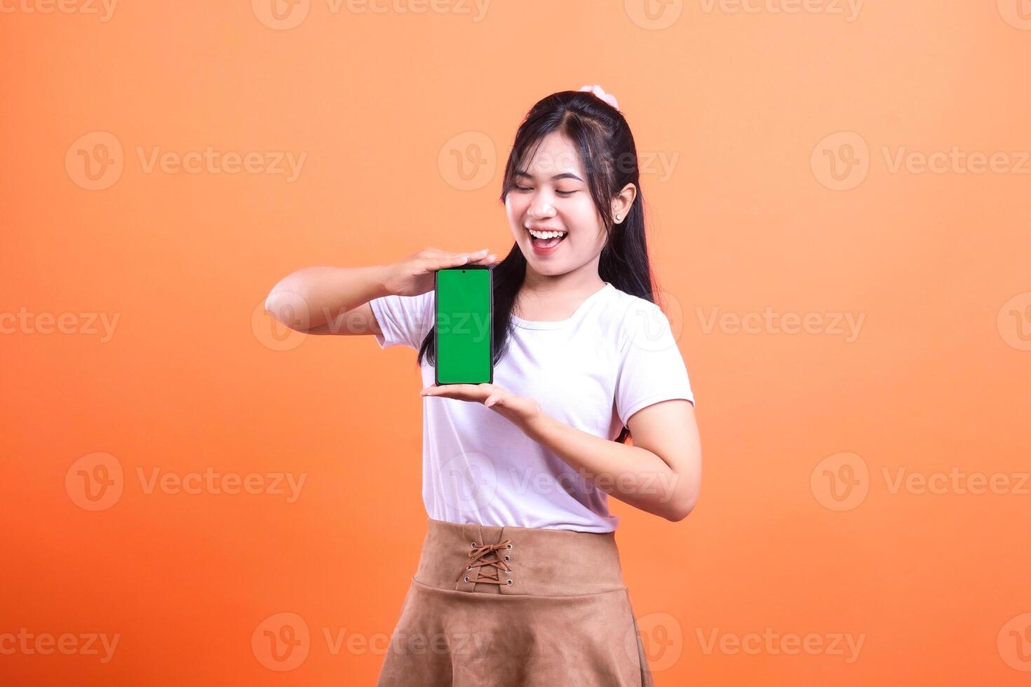 A woman is smiling while holding a smart phone device with a green screen in front of an orange background. She is wearing a white t-shirt and brown skirt. isolated on orange background photo