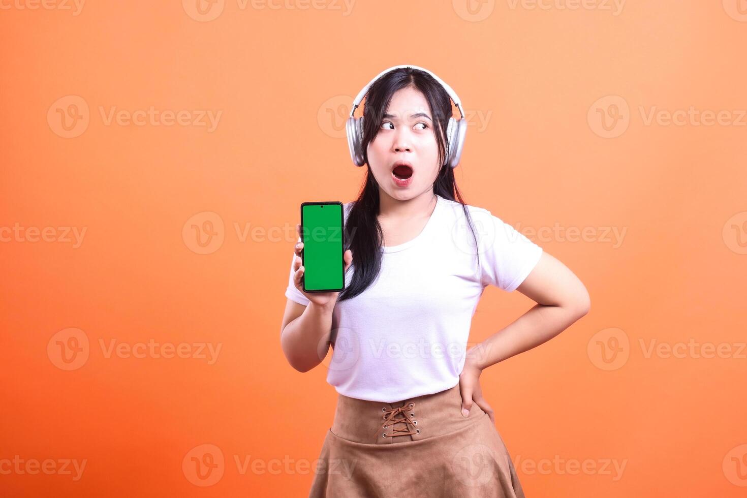 A adult woman wearing a white shirt, brown skirt and headphones holds a smart phone device gadget with a green screen in her hands and looks to her side in surprise, isolated on an orange background photo