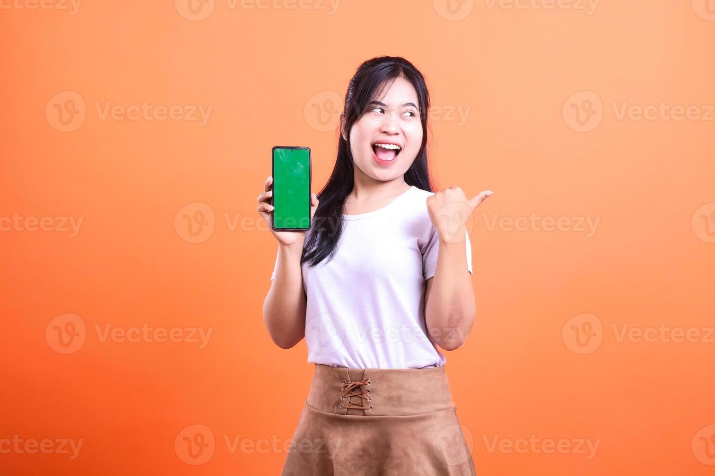 Enthusiastic woman presents smartphone with green screen on solid orange backdrop photo