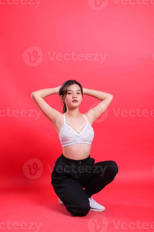 A woman in a white sports bra and black pants squats with her hands behind her head, isolated on a red background. photo