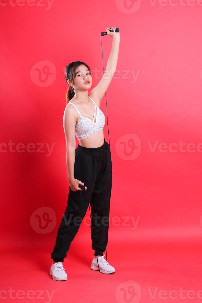A full shot of a woman holding a jump rope with one hand overhead while the other holds the jump rope handle, isolated on a red background. photo