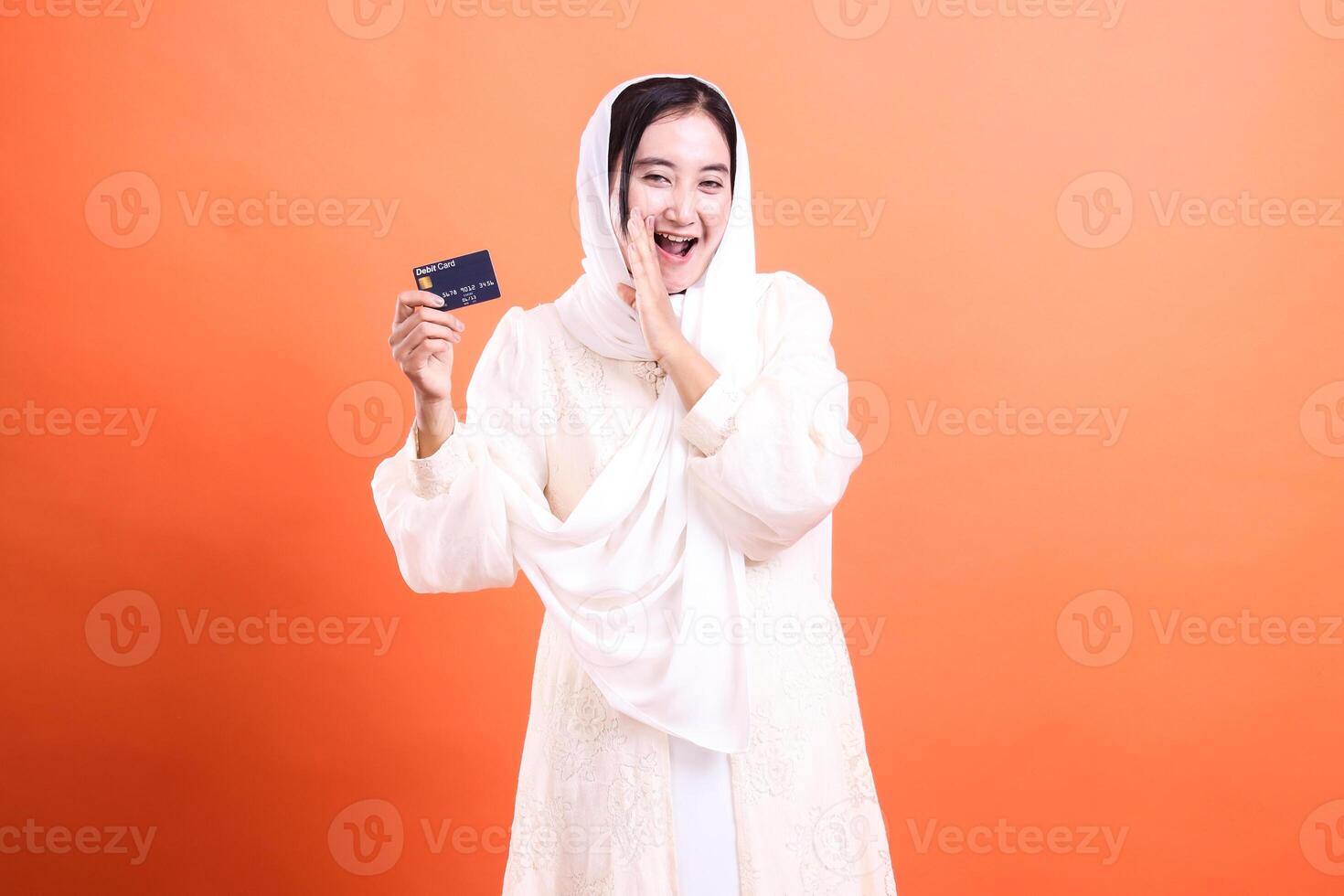 Excited Muslim Woman Holding Credit Card A sign of financial opportunity and modern convenience, isolated on a orange background. photo