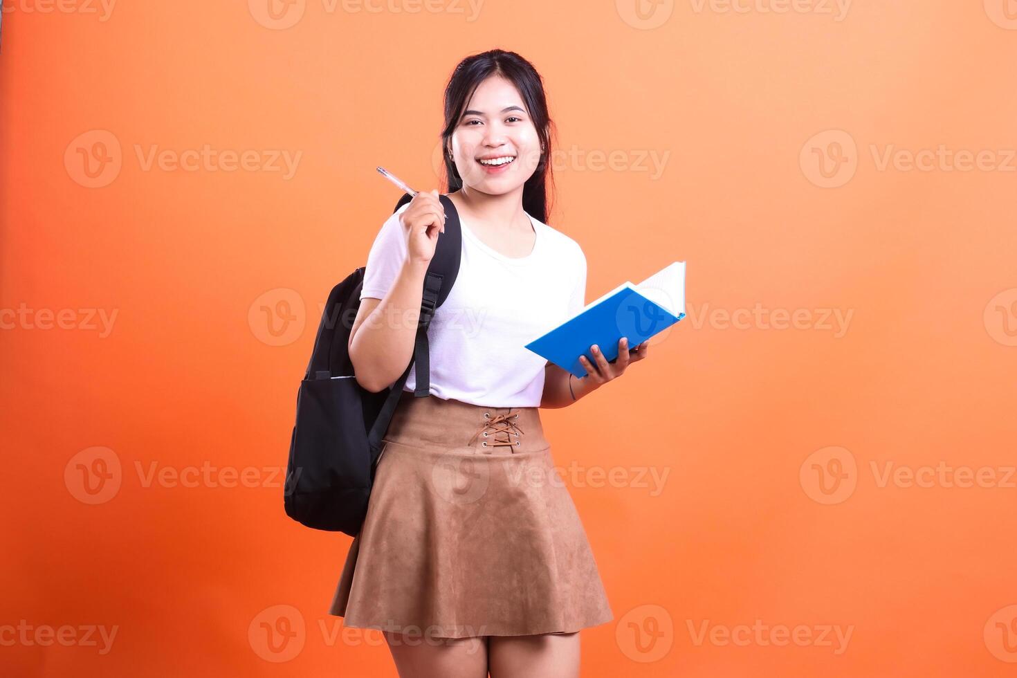 A student with a backpack and notebook holding a pen posing in front of an orange background photo