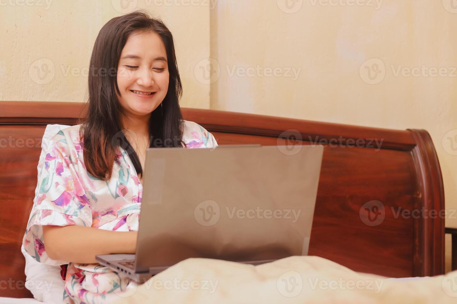 Smiling woman works from bed using her laptop, wearing a floral photo