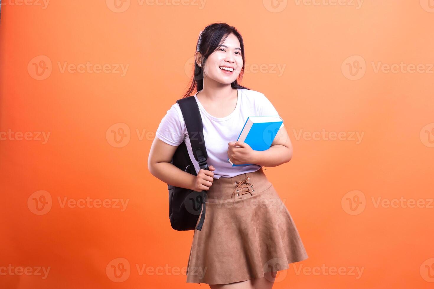 Young woman with a book and backpack, cheerfully looks away isolated on orange background photo