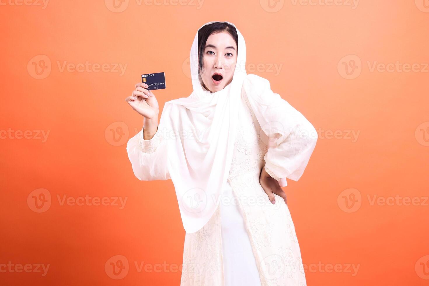 Surprised Muslim woman holding credit card with shocked facial expression, showcasing modern banking solutions and online shopping, isolated on a orange background. photo
