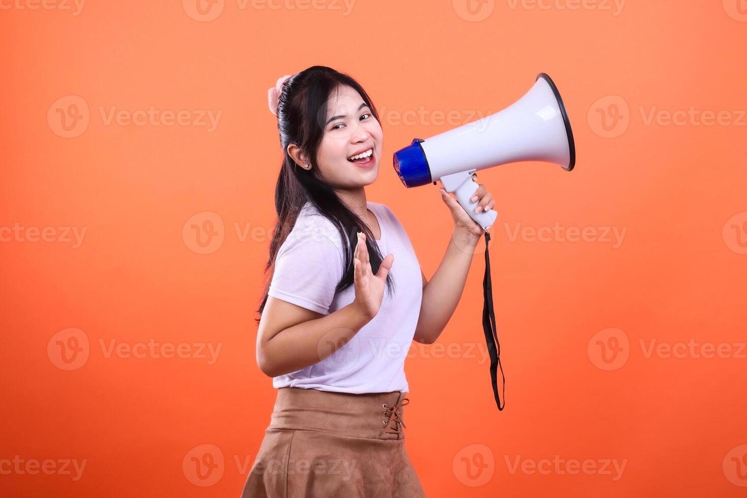 A woman shouts an announcement in a white shirt and brown skirt holding a megaphone and waving her arms against an orange background. photo