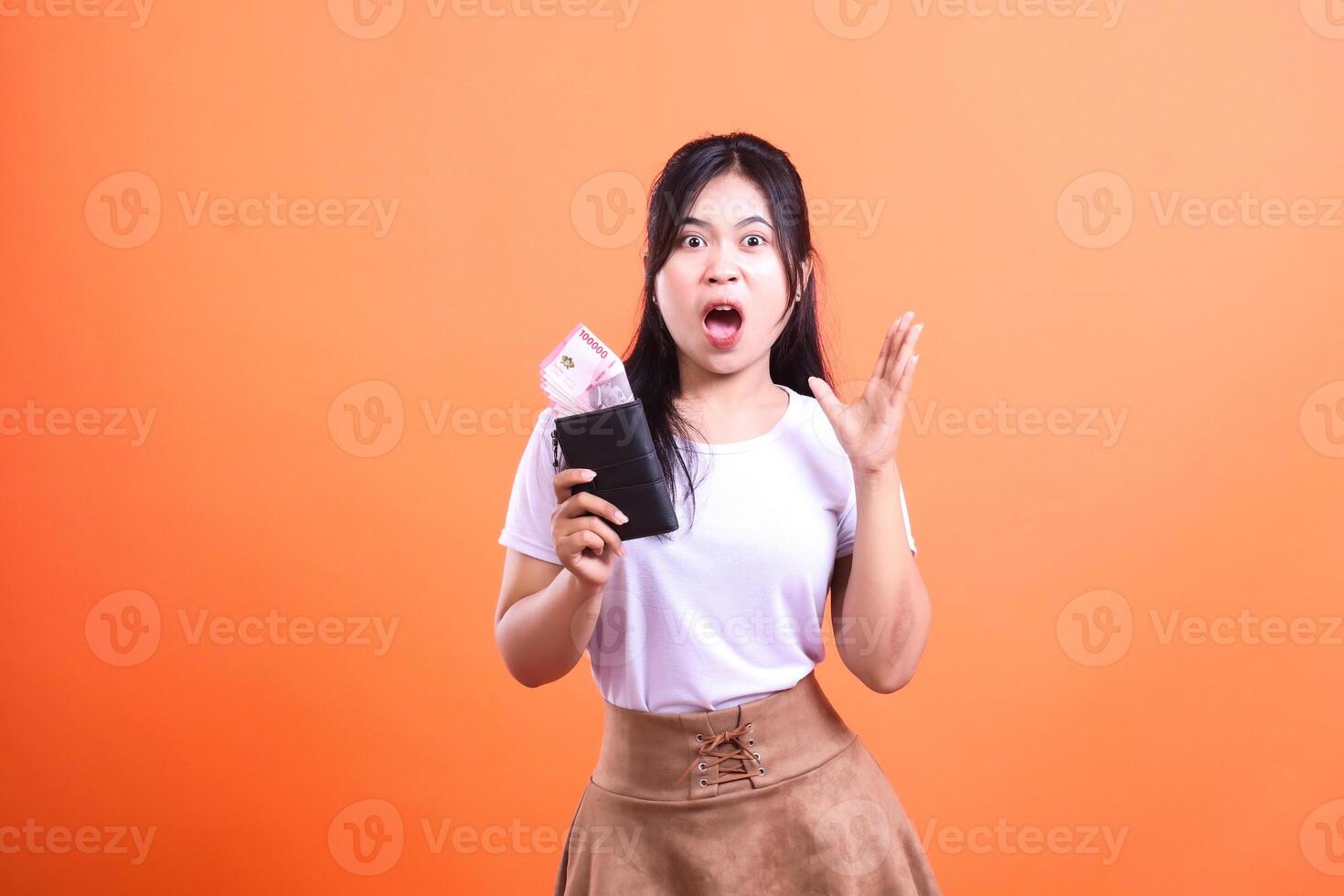 A surprised woman holding a wallet with money in hand on orange background photo