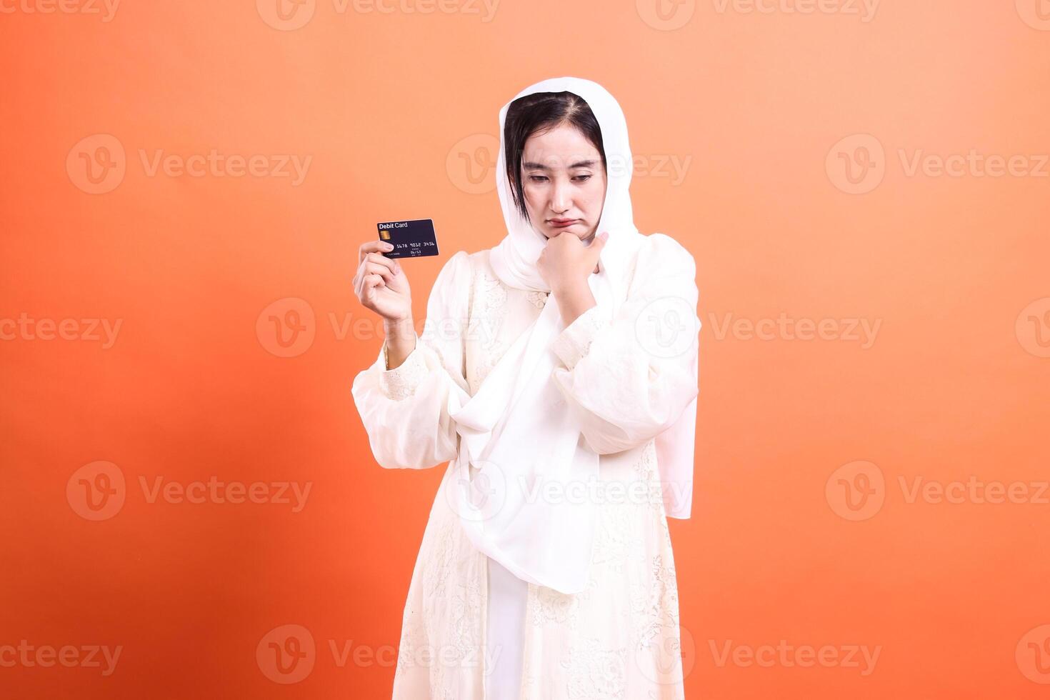 Worried Muslim Woman with Credit Card Financial Concerns Expressed, isolated on a orange background, illustrating consumer issues and banking challenges. photo