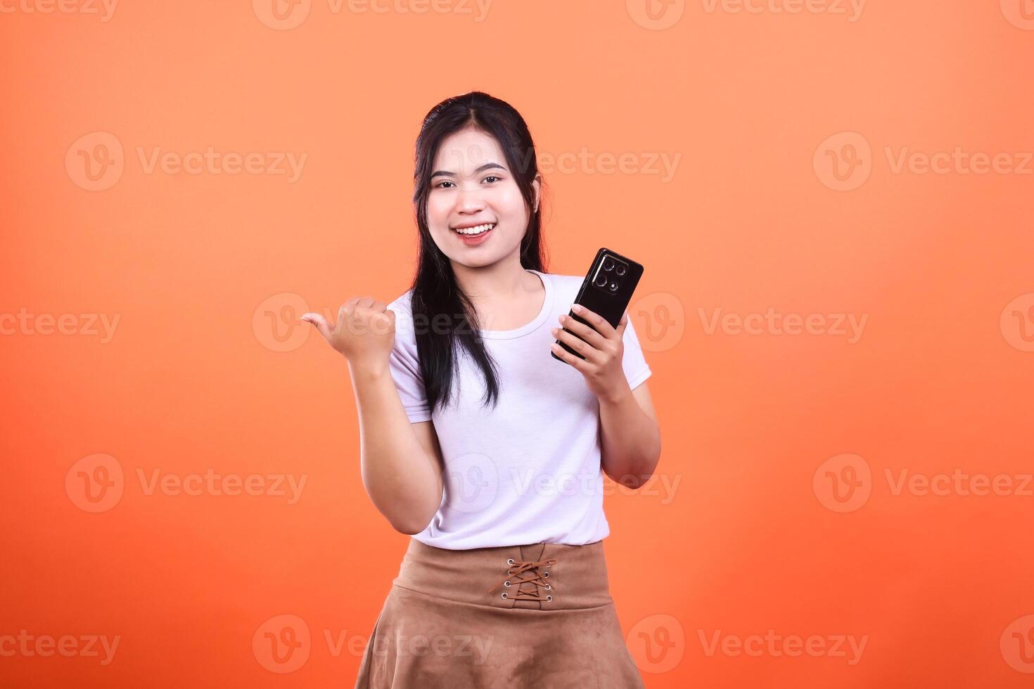 Woman holding a phone and pointing on orange background, isolated on orange backdrop photo