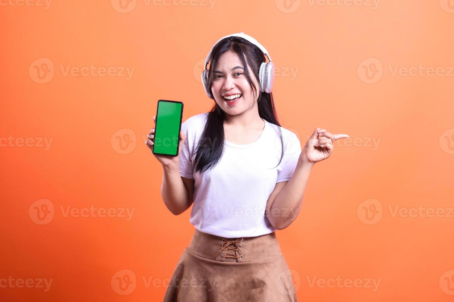 A woman with headphones holding a phone with a green screen against an orange background photo
