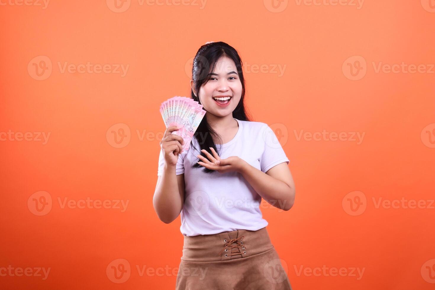 A smiling woman with a fan of currency bills, standing on an orange backdrop photo