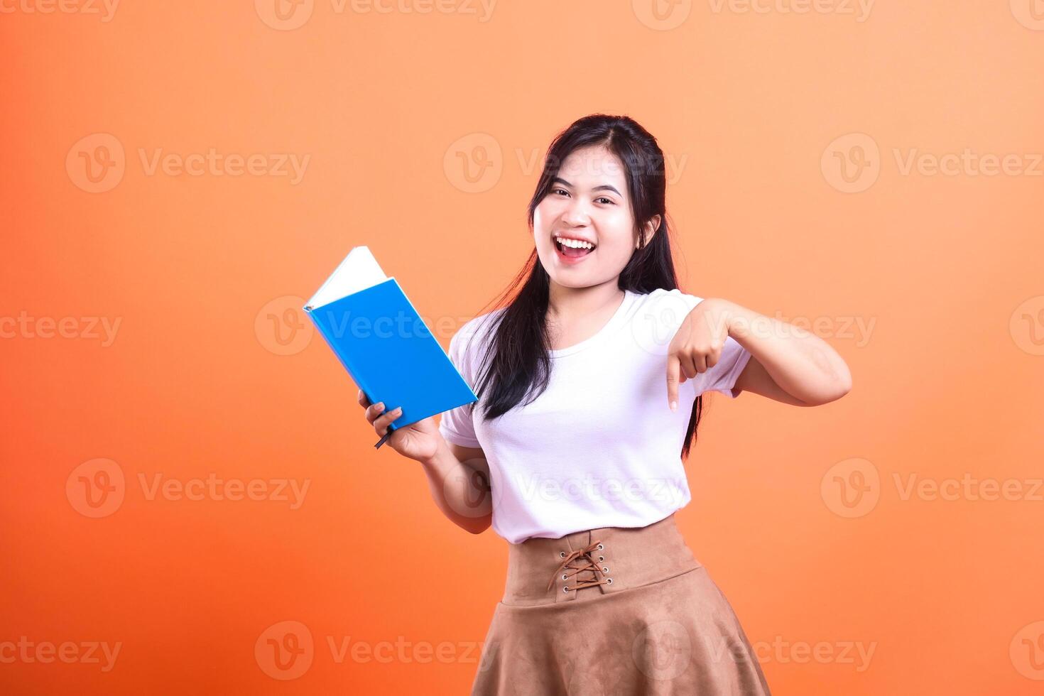 Young woman in white shirt and brown skirt holding a book and pointing down isolated on orange background photo