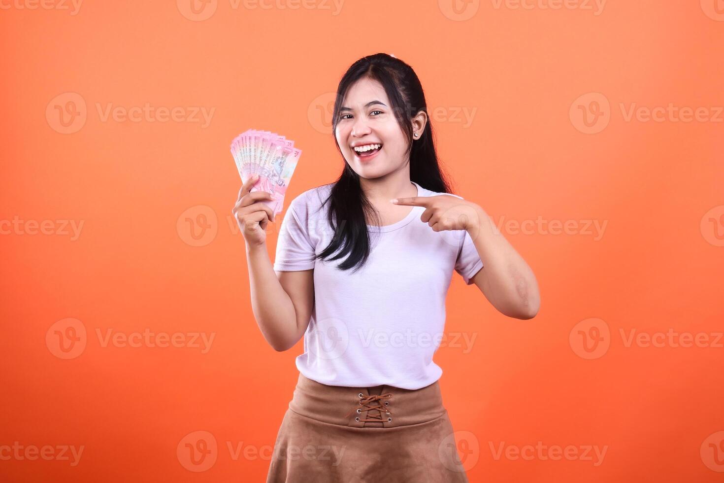 A young woman points to a pile of money, standing in front of an orange backdrop photo