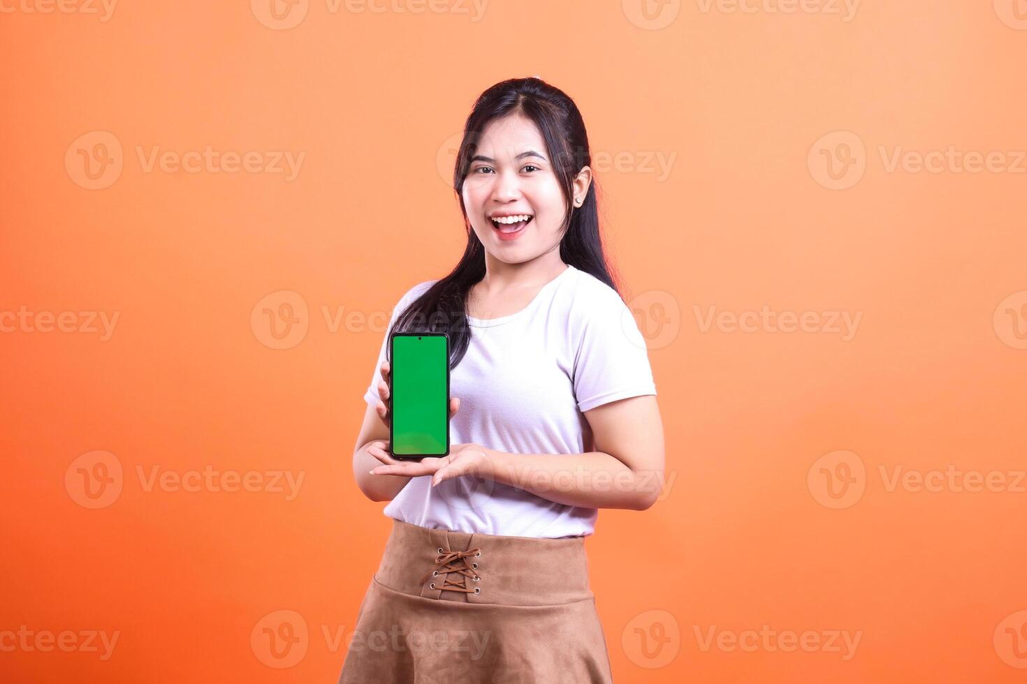 A woman holding a smart phone gadget with a green screen smiling at the camera on an orange background photo