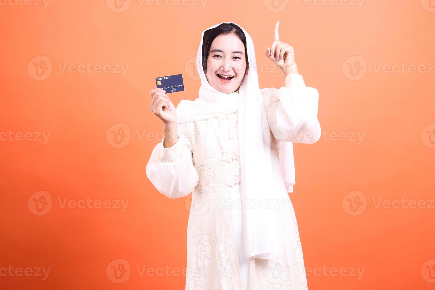 Excited Woman in Hijab Holding Credit Card and Pointing Upward, A Modern Take on Islamic Finance and Opportunity, isolated on a orange background. photo