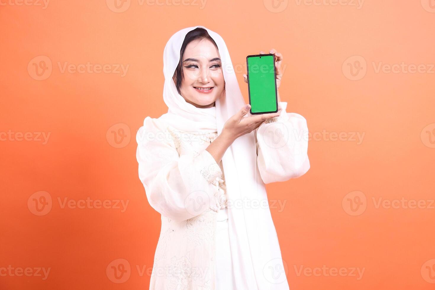 Smiling Muslim woman in hijab presents a smartphone with green screen display, happy to communicate to the user. isolated on a orange background photo