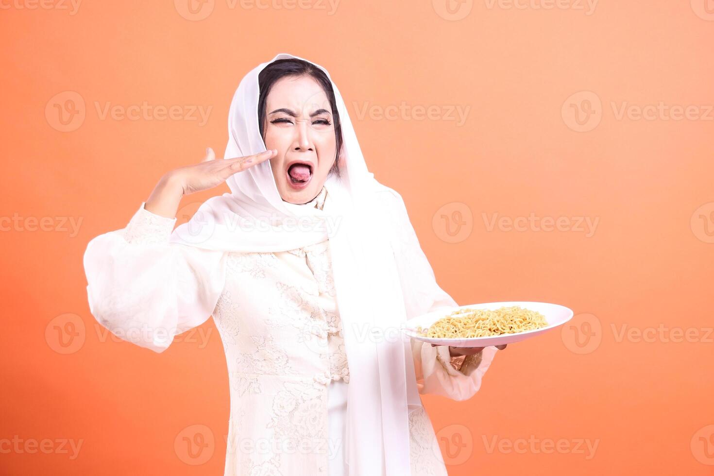 Muslim woman in traditional dress expressing disgust and aversion to noodles held on a plate in her hand, isolated on a orange background. photo