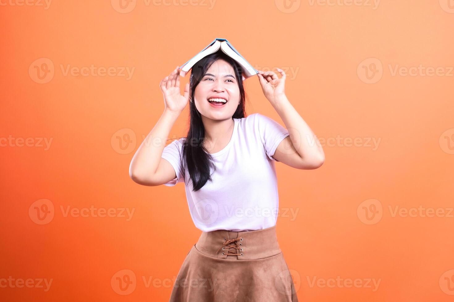A woman with a white shirt and a brown skirt has a book on her head in front of orange background photo