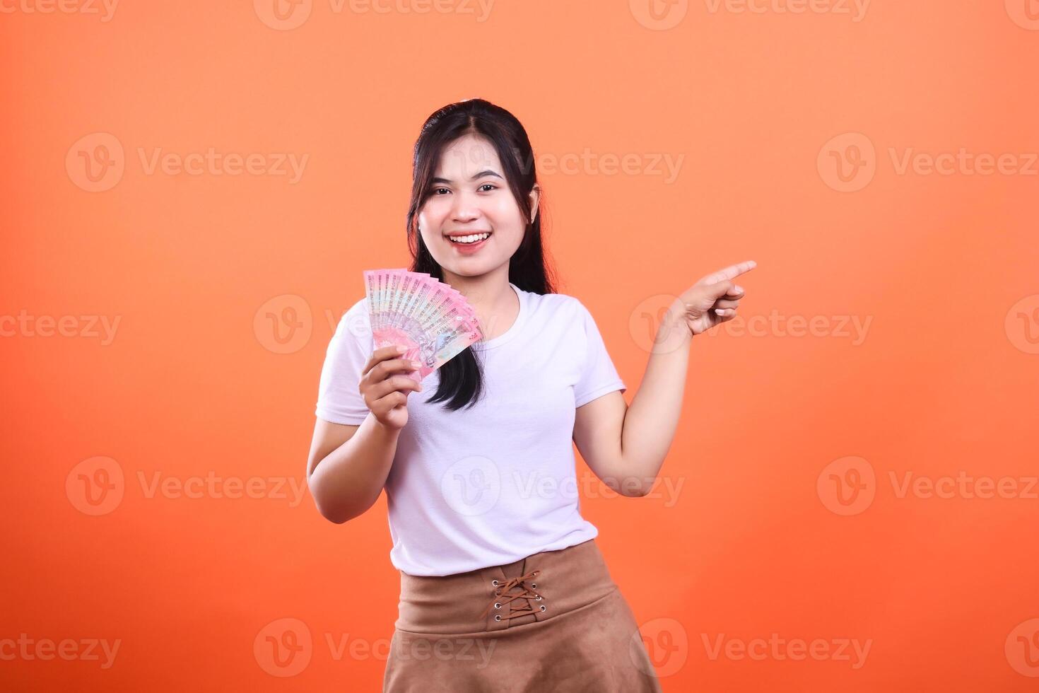 Woman holding Indonesian Rupiah bills, pointing to the side, on an orange background photo