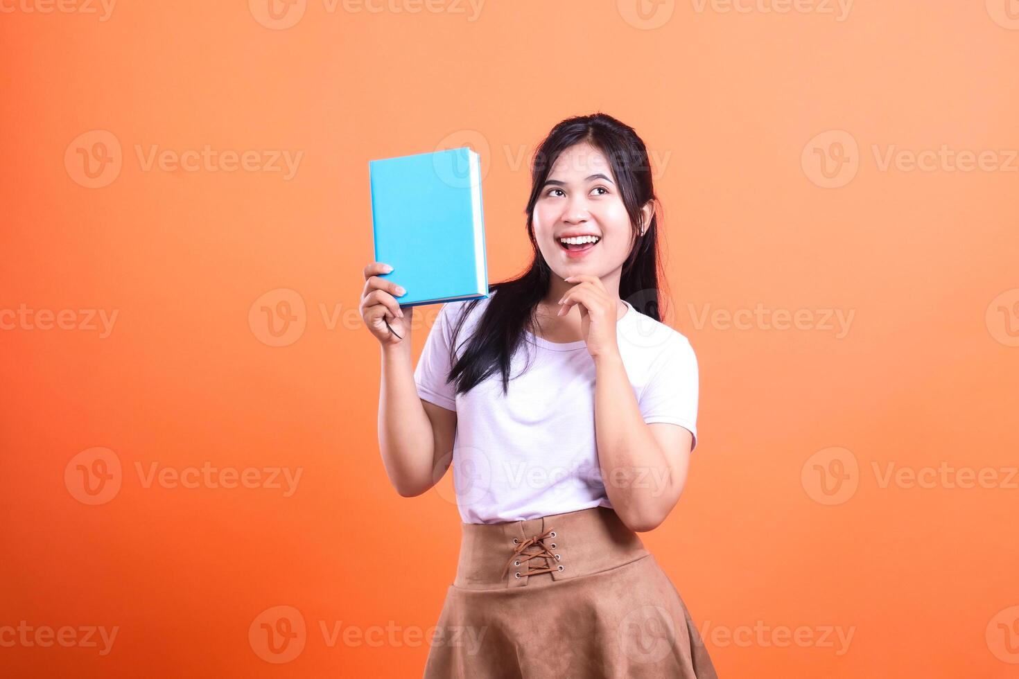 Young woman with long black hair holding a light blue book, she is wearing a white t-shirt and a brown skirt photo