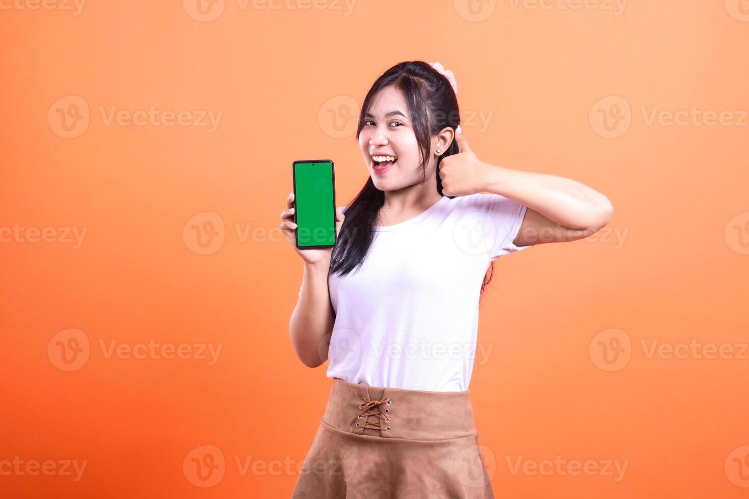 A cheerful woman is holding a smart phone gadget with a green screen and gives a thumbs up. isolated on orange background photo