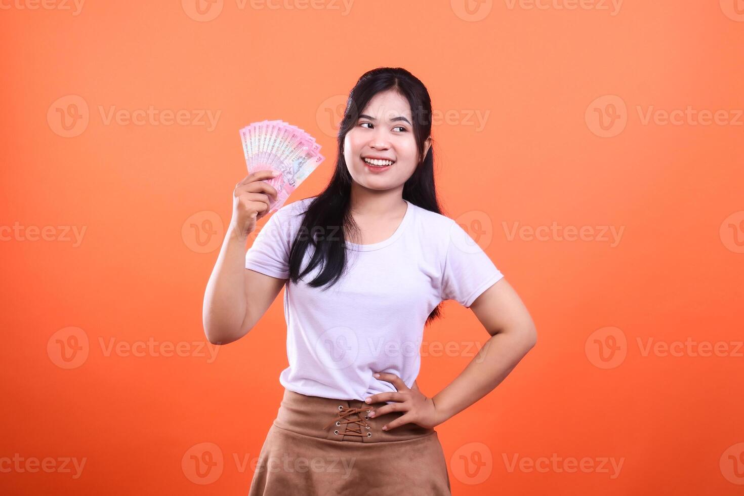 A woman holding a fan of pink paper money isolated on an orange background smiling slightly looking away photo