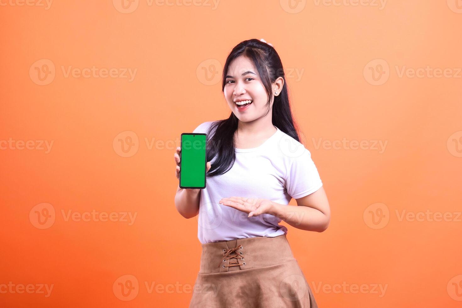 Smiling woman presents mobile phone with green screen against an orange background photo