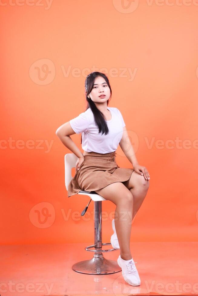 Woman in White Top and Brown Skirt Posing on a Bar Stool against Orange Backdrop photo