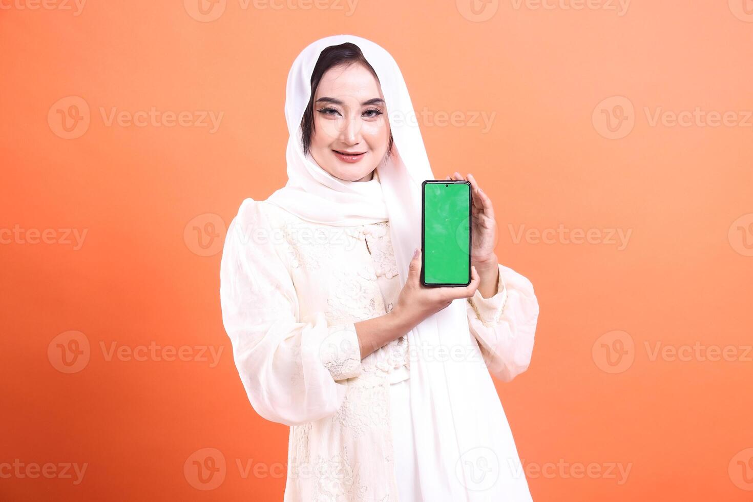 A beautiful muslim woman wearing hijab is showcasing a smartphone with a green screen, and smiling. isolated on a orange background. photo