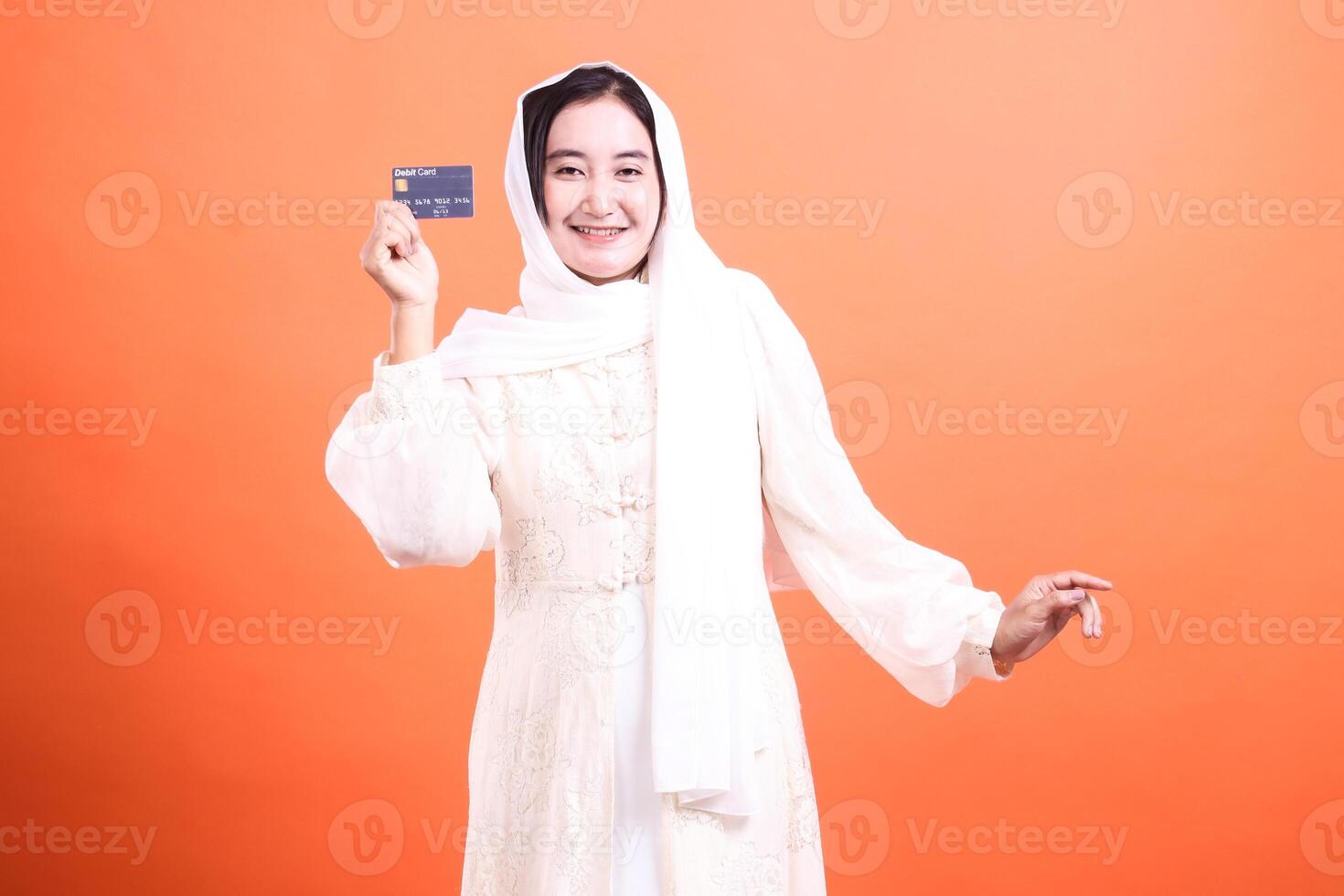Happy Muslim Woman Holding a Debit Card Smiling Positively, promoting financial services and banking options for customers isolated on a orange background. photo