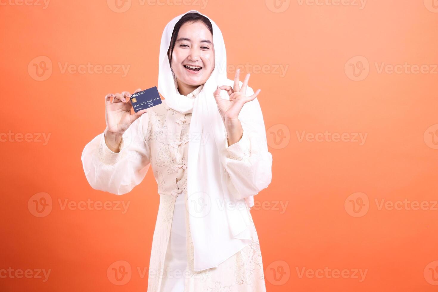 Happy Muslim Woman Holding Credit Card Showing Okay Sign, radiating positive energy and financial confidence with a smile, isolated on a orange background. photo