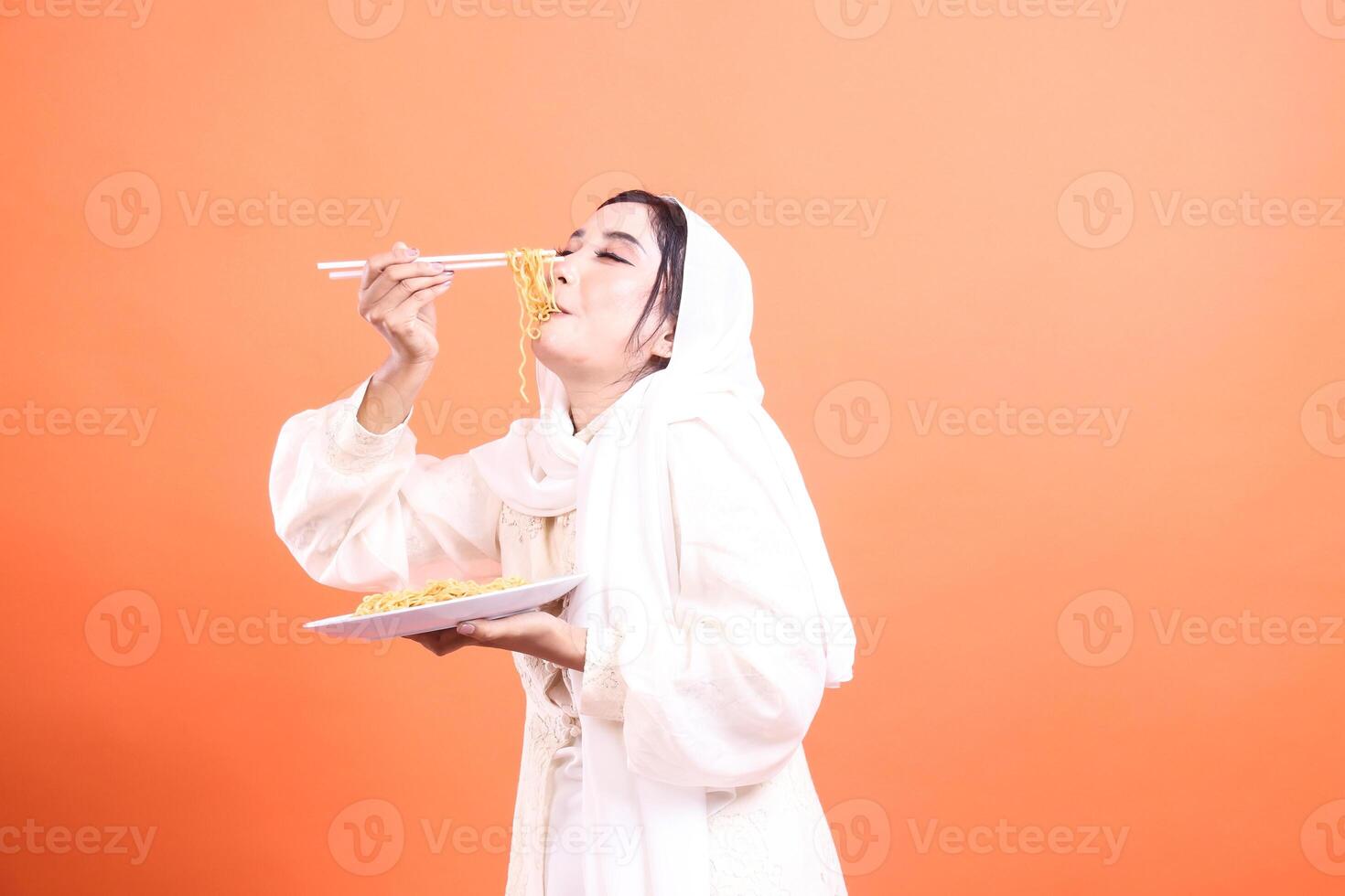 Beautiful young Muslim woman in hijab eating delicious noodles with chopsticks. Traditional Asian food in modern lifestyle isolated on a orange background. photo