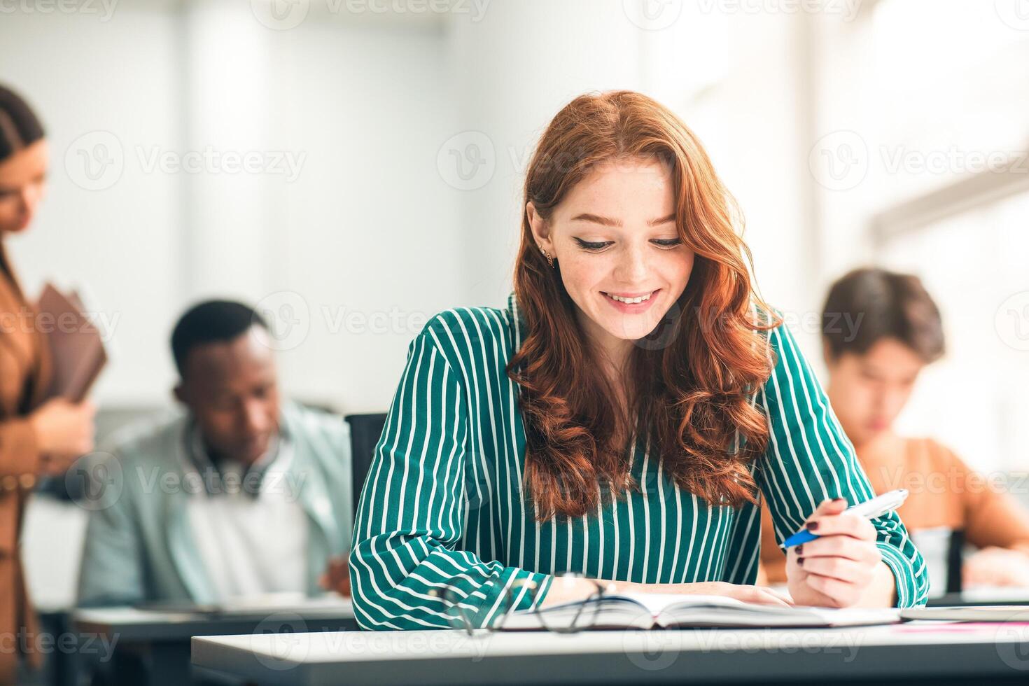 New Highschool Year On Campus. Positive Beautiful Young Adolescent Redhead Lady Sitting At Table In Modern Classroom, Doing Test Exercise, Reading Task, Enjoying Education And Learning With Classmates photo