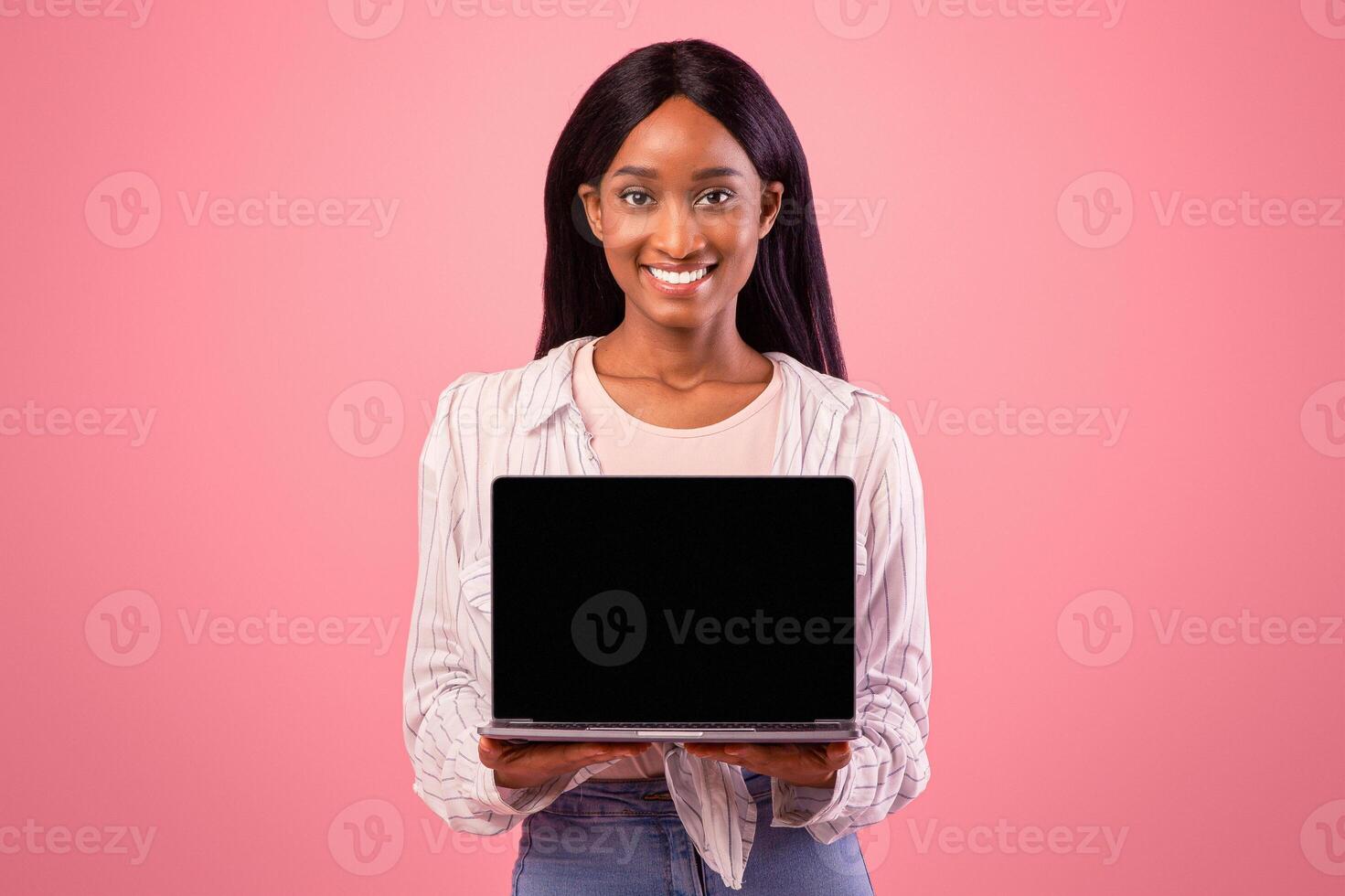 Smiling African American woman showing laptop with empty screen on pink studio background, mockup for website design. Lovely black lady with modern gadget, blank space. Online life concept photo
