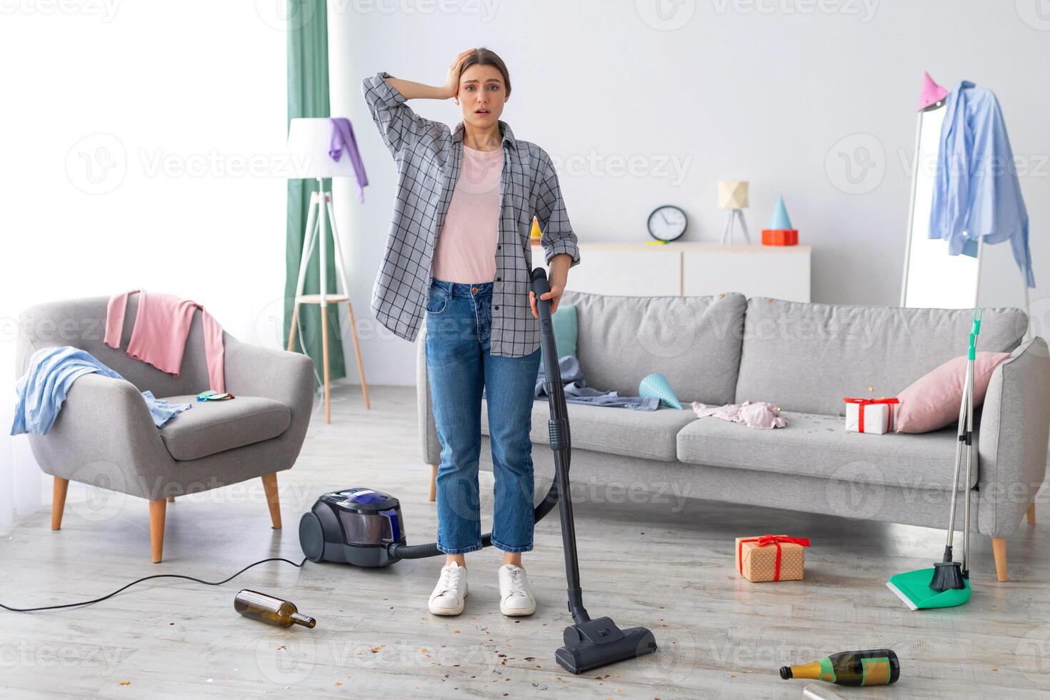 After party chaos. Dismayed millennial woman standing in her messy apartment with vacuum cleaner, holding head in terror, copy space. Terrified female student cleaning dirty university dorm room photo