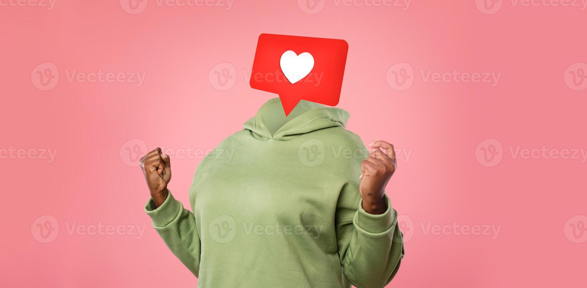 A person in a green hoodie expresses excitement and joy as a heart icon hovers instead of their head. Background is a soft pink, emphasizing the emotion linked to social media interactions and likes. photo