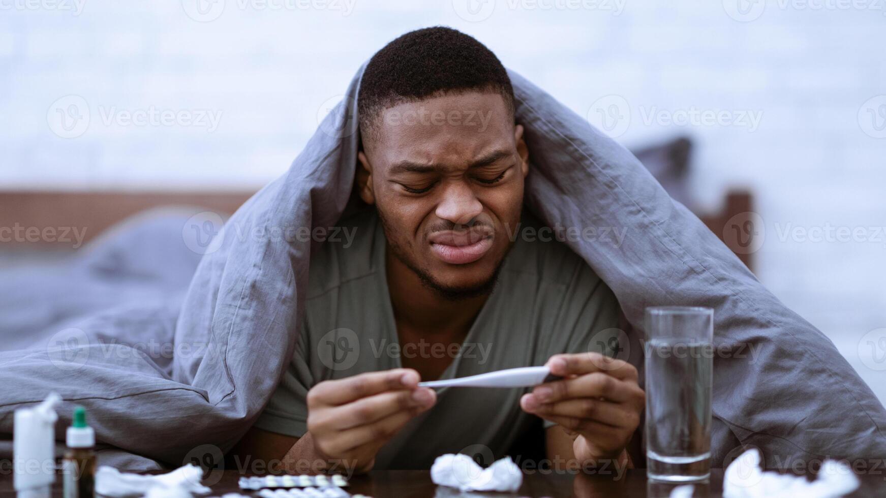 Sick Black Man Holding Thermometer Having Fever Symptom Lying In Bed At Home. African Guy Having Flu Measuring Body Temperature. Covid-19, Coronavirus And Influenza Illness. Selective Focus photo