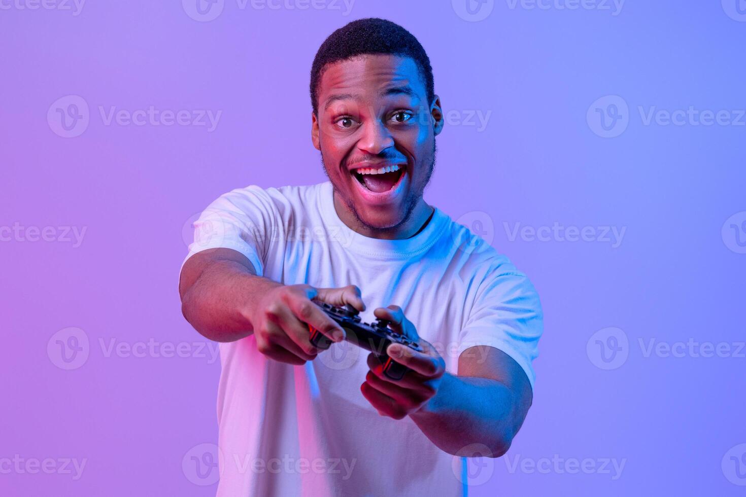 Overjoyed Black Gamer Man With Joystick Playing Games In Neon Lighting, Emotional Excited Guy Having Fun While Standing In Neon Lighting Over Purple Studio Background, Looking At Camera photo