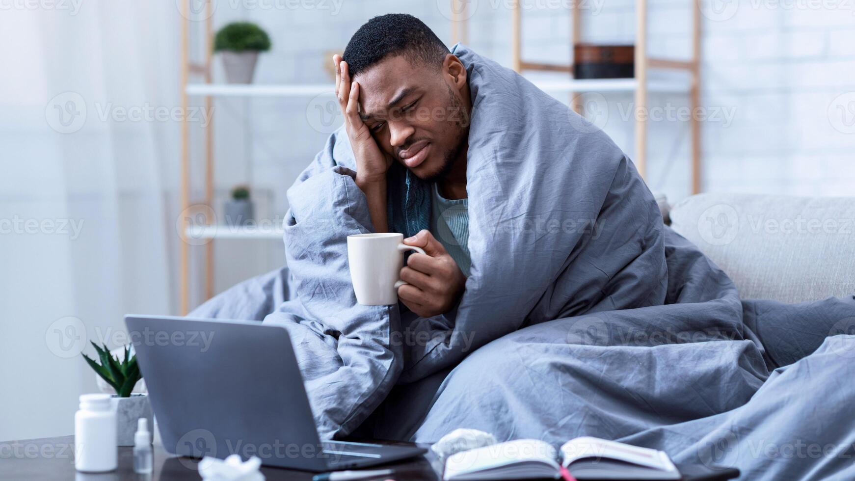Sick African American Man Using Laptop Working While Being Ill, Having Fever And Headache Sitting On Sofa Wrapped In Blanket At Home. Guy Browsing Internet For Influenza, Cold, And Covid-19 Symptoms photo