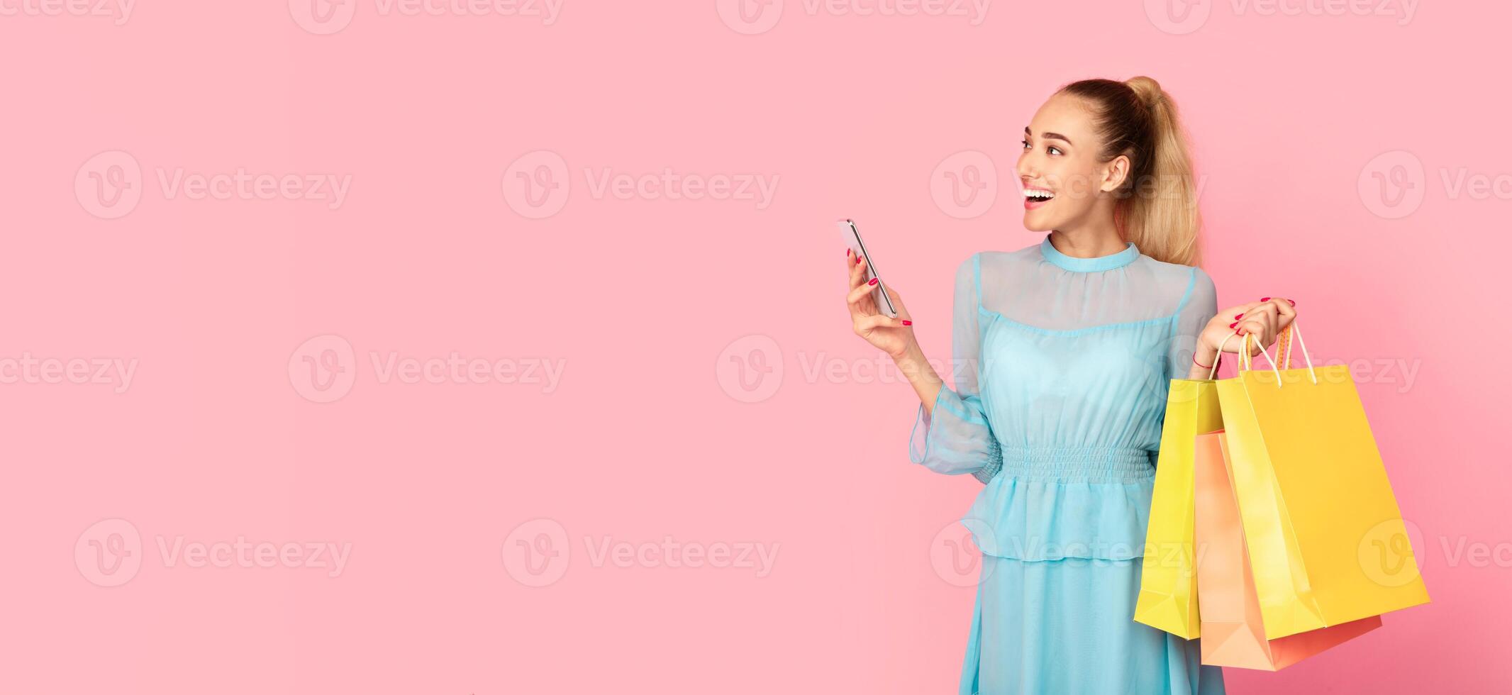 Portrait of smiling young woman in dress carrying colorful shopping bags, holding smartphone and looking at copy space for advert, text or promo, standing isolated on pink studio background photo