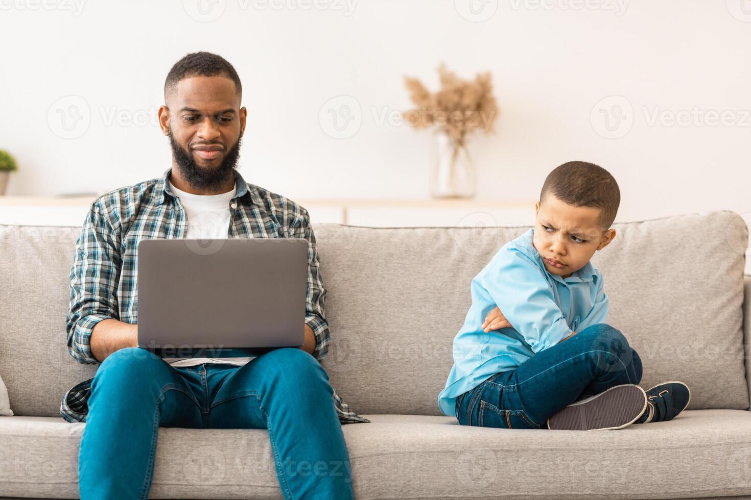 Father's Indifference. Black Dad Ignoring Offended Son Using Laptop Sitting On Couch Indoors. Reluctant Daddy Neglecting His Unhappy Kid. Bad Relationship With Father Concept photo
