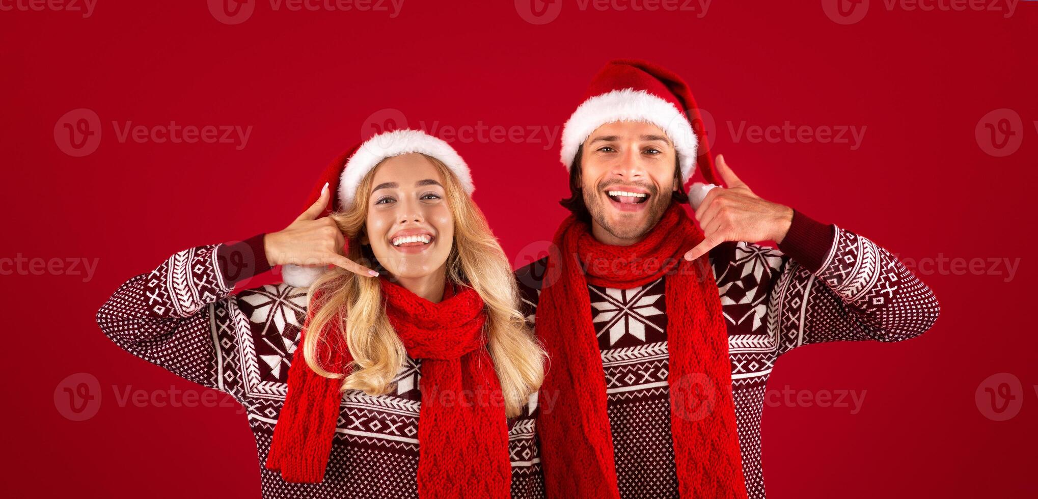 Joyful young couple in Christmas sweaters and Santa hats showing CALL ME gesture on red background photo