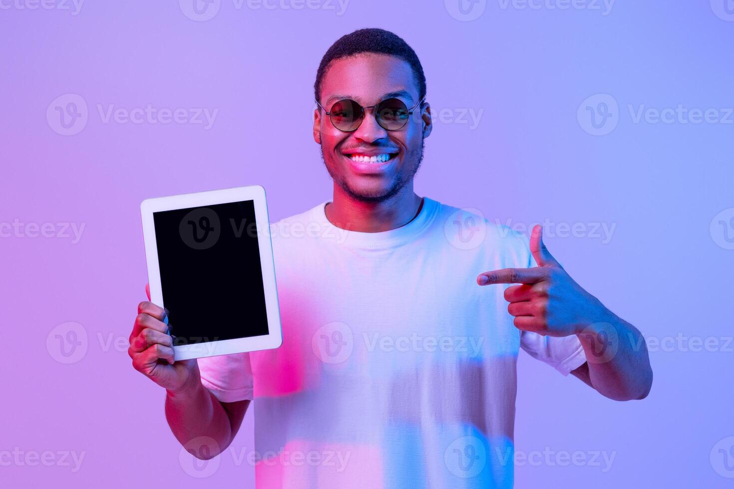 Cheerful Black Man Pointing At Blank Digital Tablet Screen, Demonstrating Copy Space For Your App Or Website Design, Standing Under Vivid Neon Light Over Purple Studio Background, Mockup Image photo