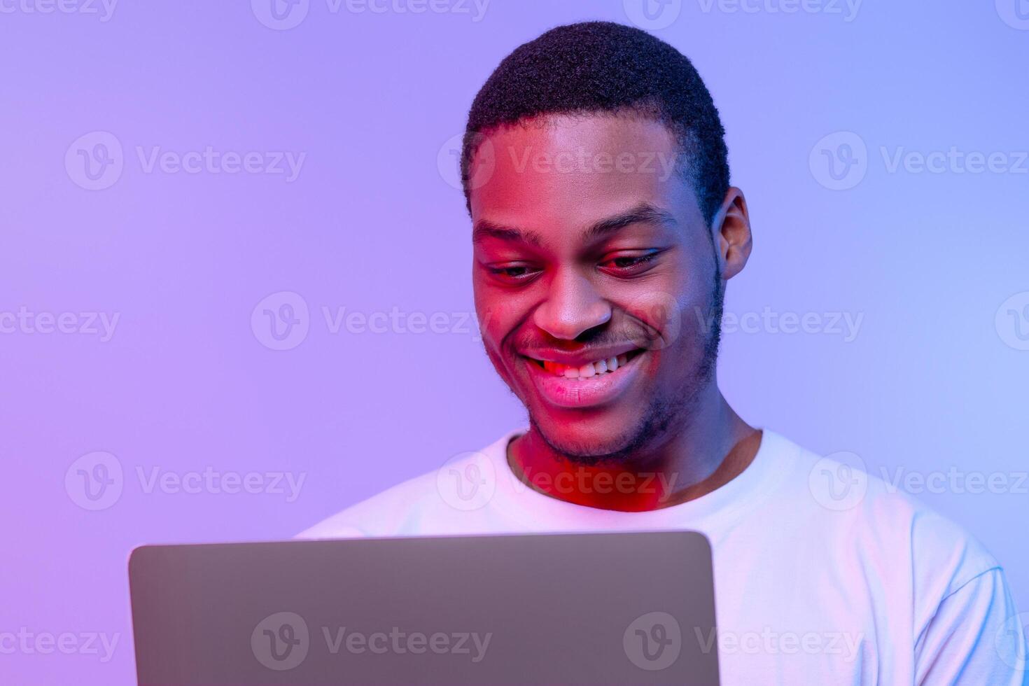 Handsome Smiling Black Programmer Guy Working Online On Laptop In Neon Light, Cheerful African American Freelancer Man Using Computer In Neon Light Over Purple Studio Background, Closeup Portrait photo