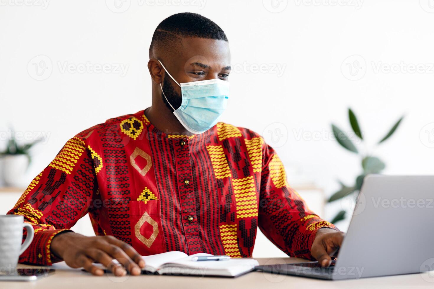 Work During Pandemic. Black Freelancer Guy In Medical Mask Using Laptop At Home, Having Remote Job During Coronavirus Lockdown, African Man In Traditional Clothes Sitting At Desk, Typing On Keyboard photo
