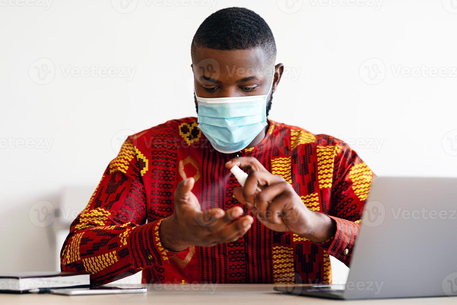 Portrait Of Young African Man Wearing Mask Sanitizing Hands While Sitting At Desk In Office, Applying Disinfectant Spray, Black Guy In traditional Clothes Keeping Protective Measures During Pandemic photo