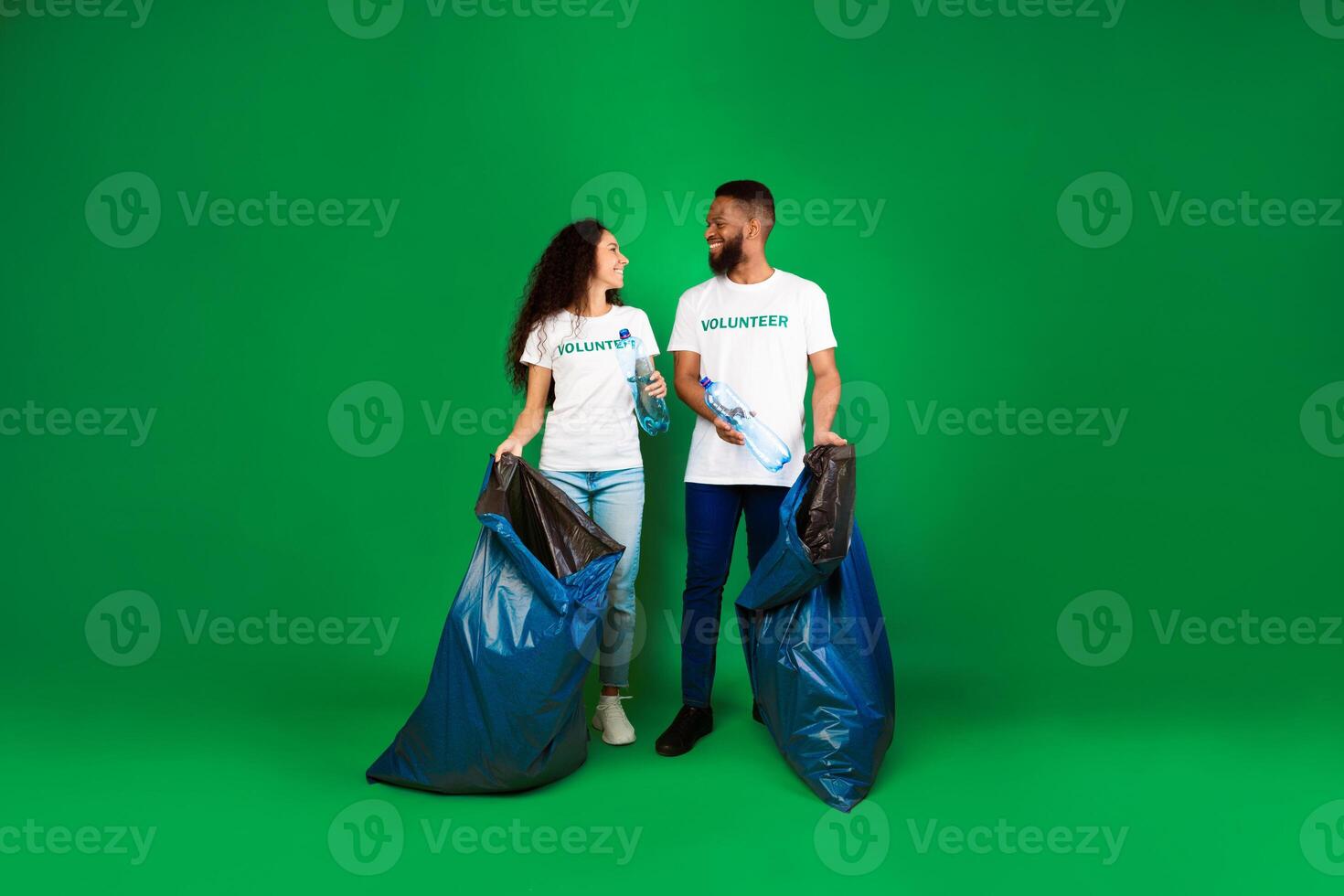 Two Diverse Volunteers With Big Blue Garbage Bags Picking Wasted Plastic Bottles Posing On Green Background. Studio Shot. Keep Environment Clean, Ecology And Volunteering Work Concept. Full Length photo