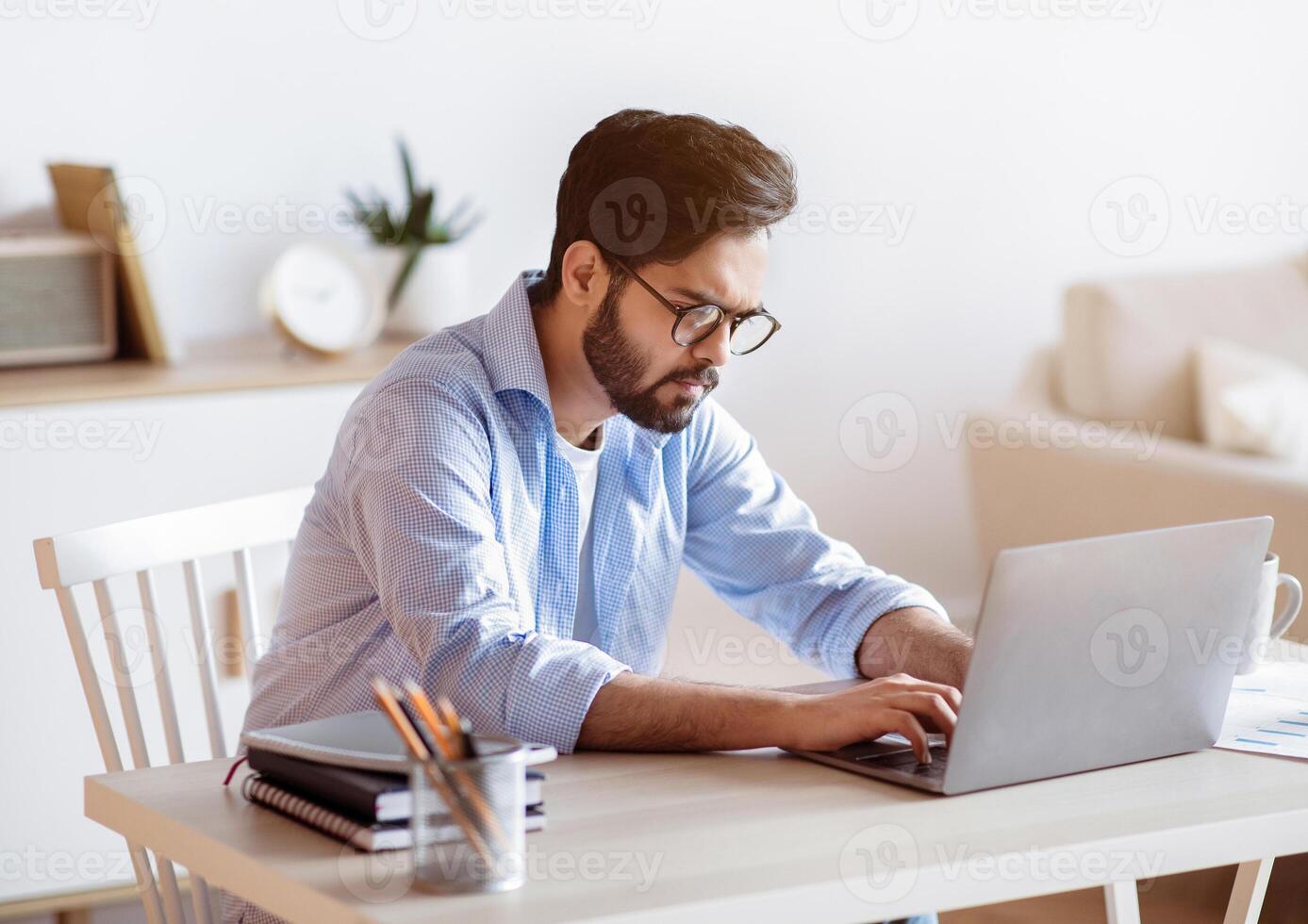 Arab Freelancer Guy Sitting At Desk At Home And Typing On Laptop. Handsome Middle Eastern Male Entrepreneur In Eyeglasses Working Online, Focused On Computer Screen, Enjoying Remote Job, Free Space photo