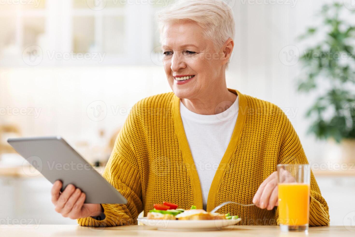 Joyful senior lady with digital tablet having lunch at home, drinking fresh orange juice, eating healthy sandwich and reading culinary blog or looking for new recipes online, free space photo